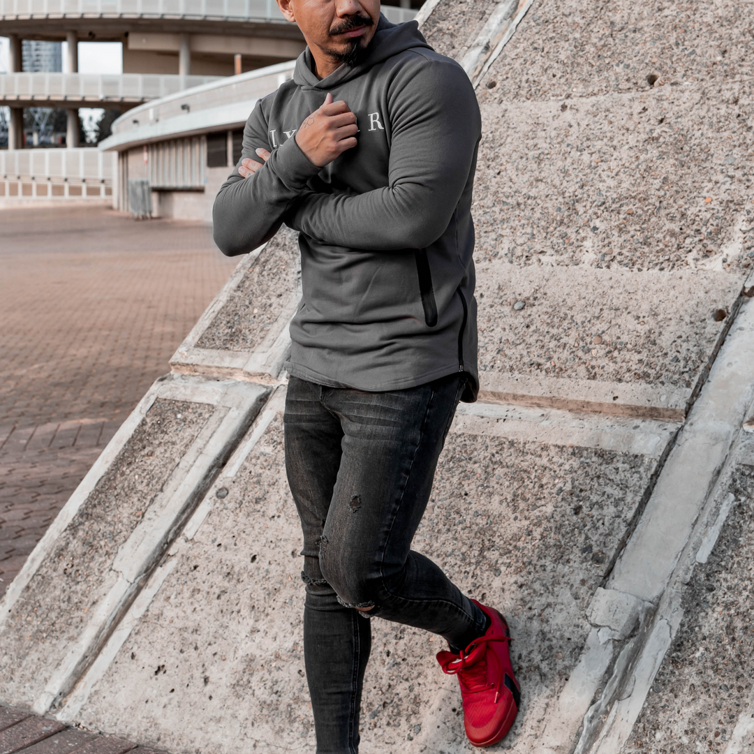 Male model is wearing the IXK Gear Muscle Hoodie in Colour: Charcoal. Concrete wall background.