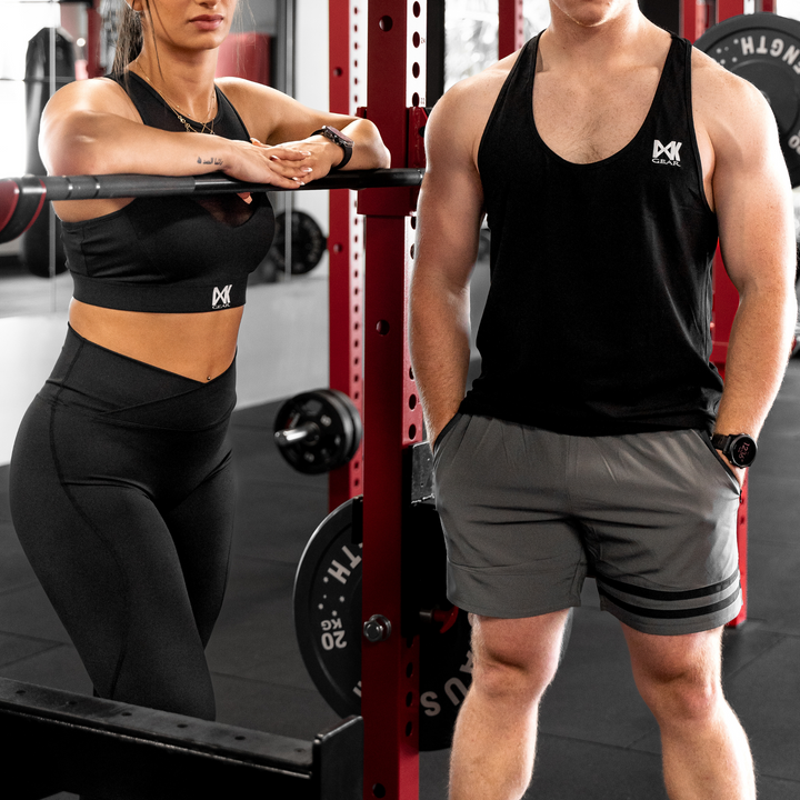IXK Gear NV Tights in Colour: Black, paired with Mesh Sports Bra in Black. Gym background. Male model is wearing the IXK Gear Stringer Tank Top in Colour: Black paired with Quick Dry Accent Shorts in Grey.