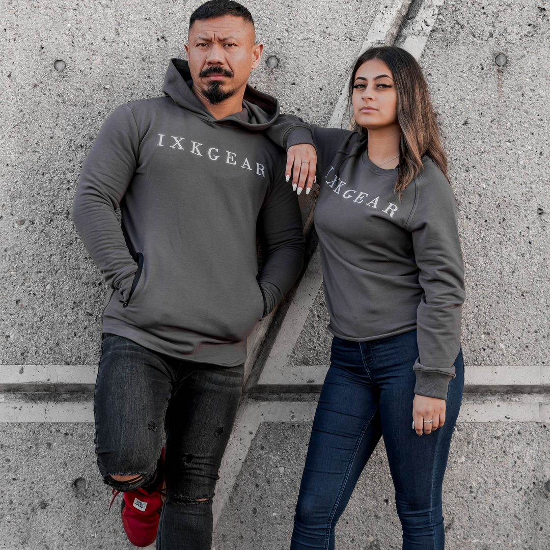 Male model is wearing the IXK Gear Muscle Hoodie in Colour: Charcoal. Concrete wall background. Female model wearing IXK Gear Crewneck Jumper in Colour: Charcoal.