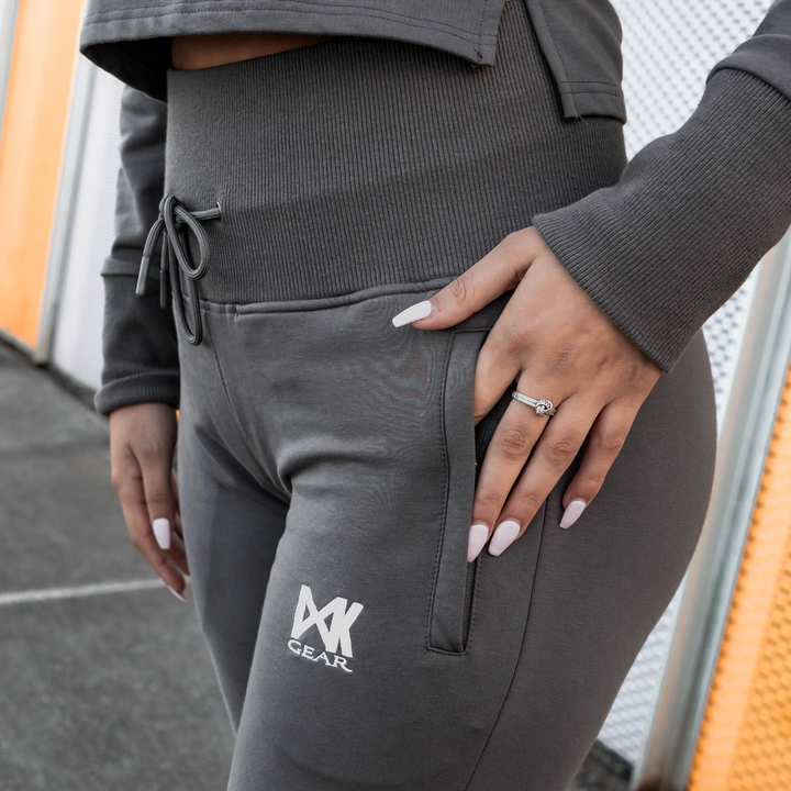 IXK Gear Women's Tracksuit in colour: Charcoal. Two-Piece tracksuit which includes slightly cropped hoodie and slim-fit trackpants trackies with zip pockets. Orange mesh background. Close p shot of the high-waisted trackpants and zip pockets.