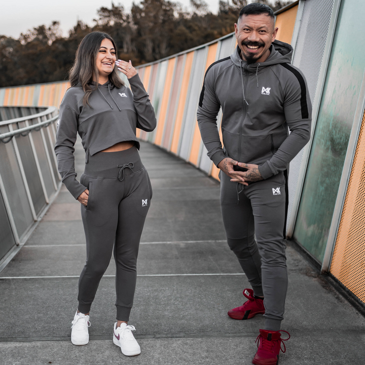 IXK Gear Women's Tracksuit in colour: Charcoal. Two-Piece tracksuit which includes slightly cropped hoodie and slim-fit trackpants trackies with zip pockets. Orange mesh background. Male model is wearing the IXK Gear Men's Tracksuit in Charcoal.