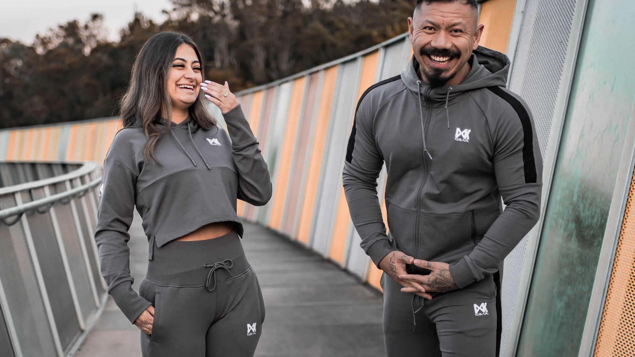 IXK Gear Women's Tracksuit in colour: Charcoal. Two-Piece tracksuit which includes slightly cropped hoodie and slim-fit trackpants trackies with zip pockets. Orange mesh background. Male model is wearing the IXK Gear Men's Tracksuit in Charcoal.