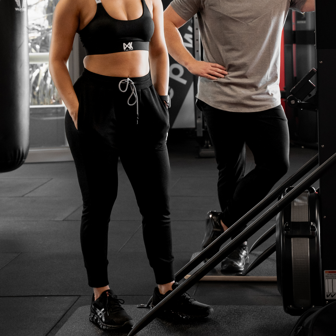IXK Gear Women's Cropped Trackies in Colour: Black paired with X sports Bra in Coloir: Black. Male model is wearing IXK Gear Cotton Tee in Grey and Slim Sweats Trackpants Trackies in Black. Gym background.