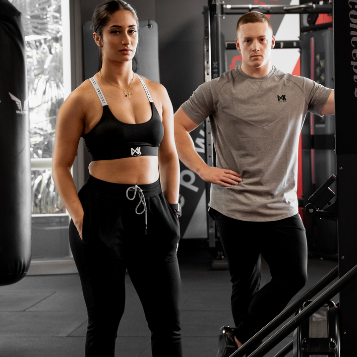 IXK Gear X Sports Bra in Colour: Black. Model is also wearing Cropped Trackies in Colour: Black. Gym Background. Male model is wearing IXK Gear Cotton Tee in Grey and Trackies in Black.